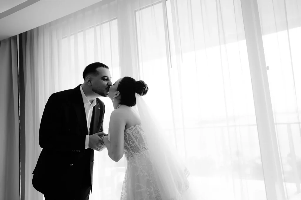 Black and white photo of the bride and groom kissing by a window